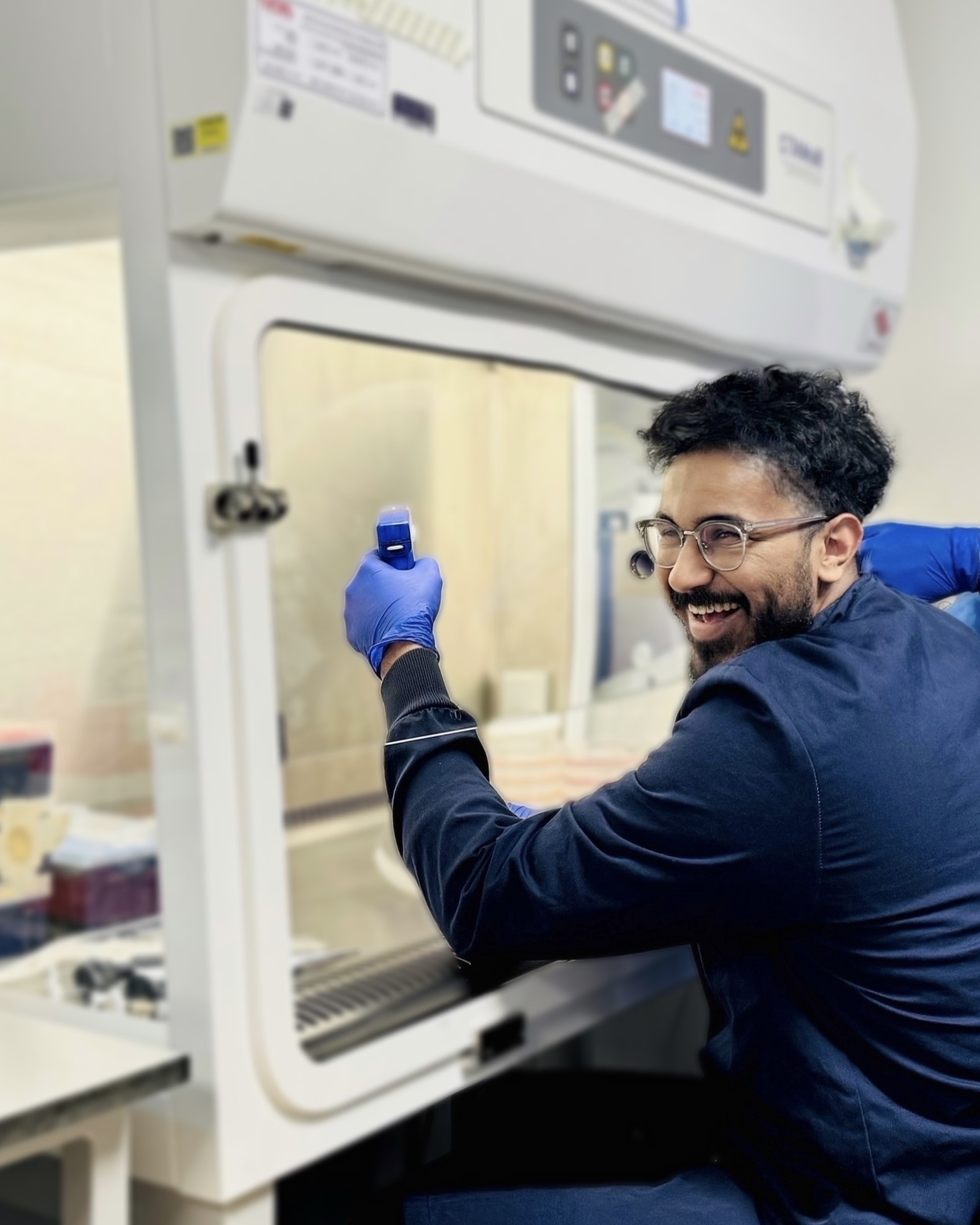 Anfas laughs whilst turning towards the camera whilst working in a tissue culture hood. He wears a blue lab coat and gloves and holds a pipette boy.